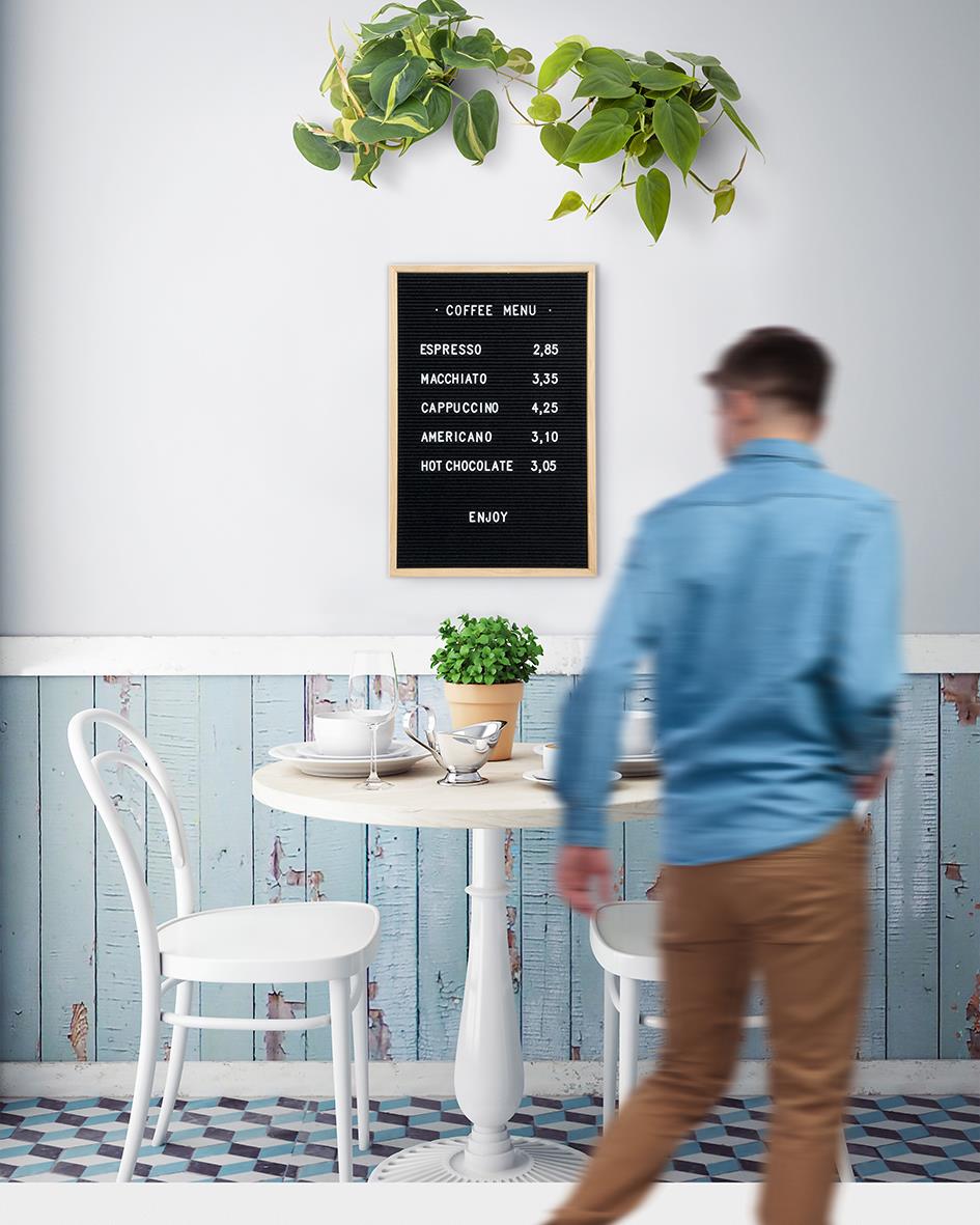 Kaffeemeny på svart anslagstavla i ett ljust café med växt och vit bord och stolar, person i blå jacka i rörelse