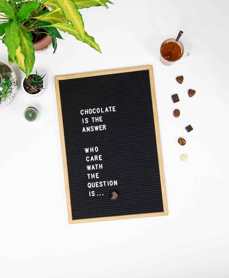 Svart bokstavstavla med citatet Chocolate is the answer, omgiven av växter, chokladbitar och ett glas te på en vit bakgrund
