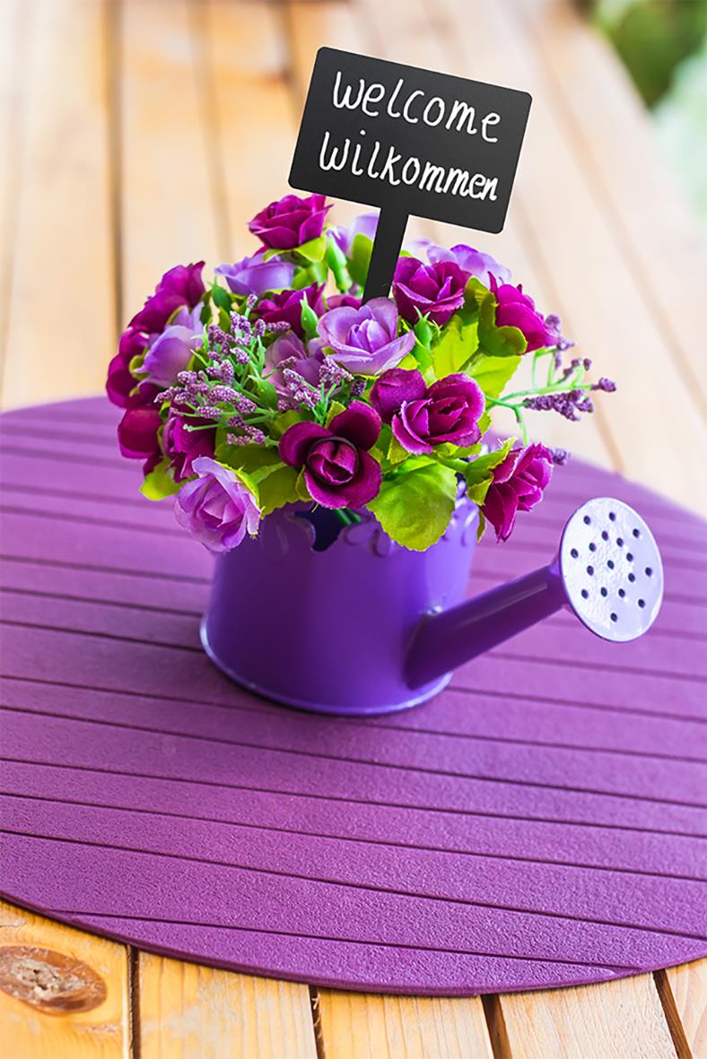 Svart rektangulär skylt med texten welcome och Willkommen i blomkruka med blommor på ett bord