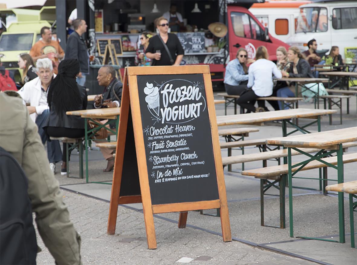 Utomhusmarknad med folk och en svart anslagstavla med text om frozen yoghurt