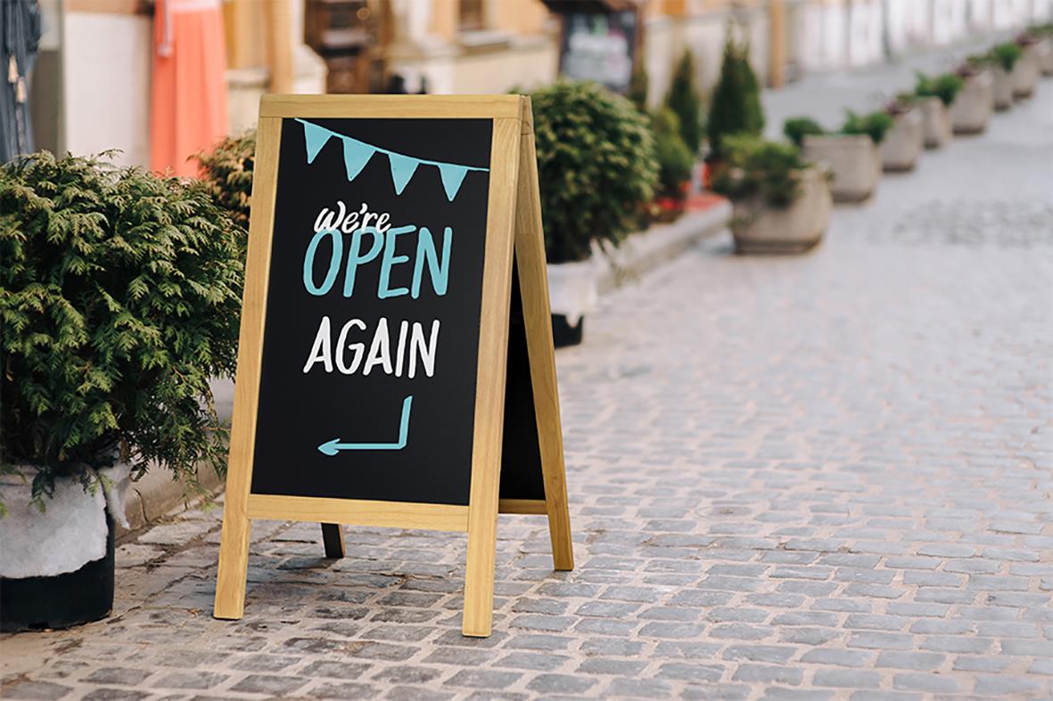 Gatuprydnadsskylt av trä med svart skrivyta och text We’re OPEN AGAIN, placerad på gatukant med växter i bakgrunden