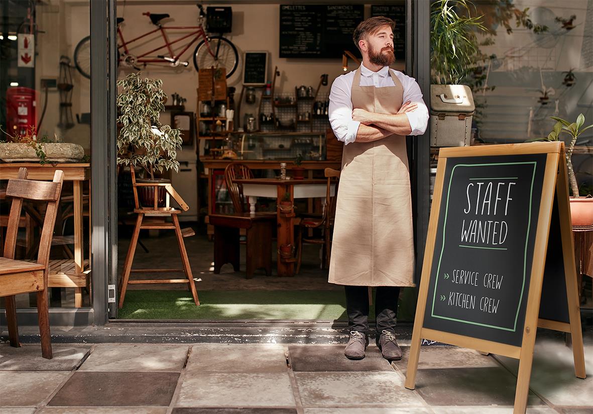 Person står utanför café med skylt som säger 'Staff Wanted' och 'Service Crew'