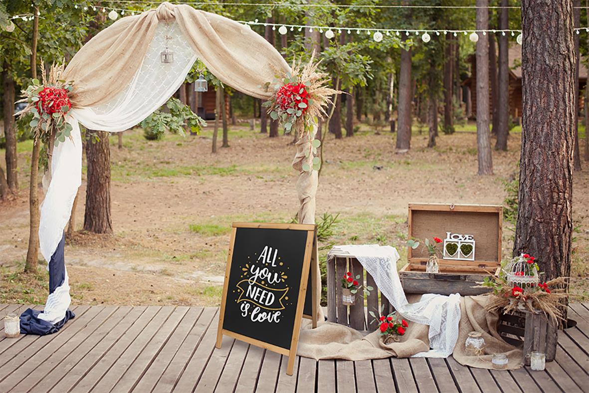 Utomhus scen med tygdraperi, blomster, svart anslagstavla och träskrin på en träplattform i skogsmiljö