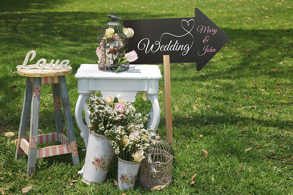 Svart griffelskylt med pil och text för bröllop, omgiven av blommor och dekorativa föremål på gräsmatta