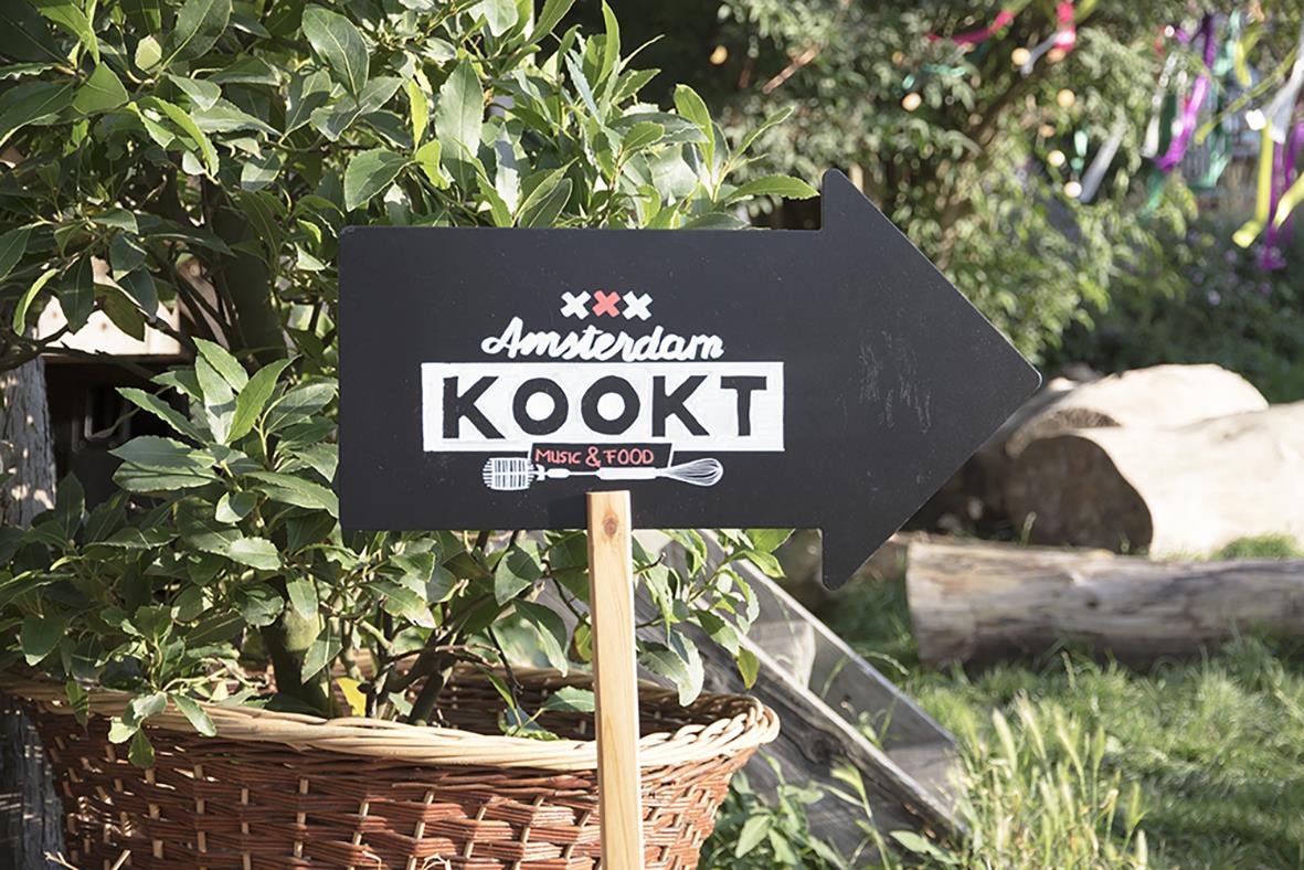 Svart skylt med pil, texten Amsterdam KOKT och en pinne, utomhus med växter och sten i bakgrunden