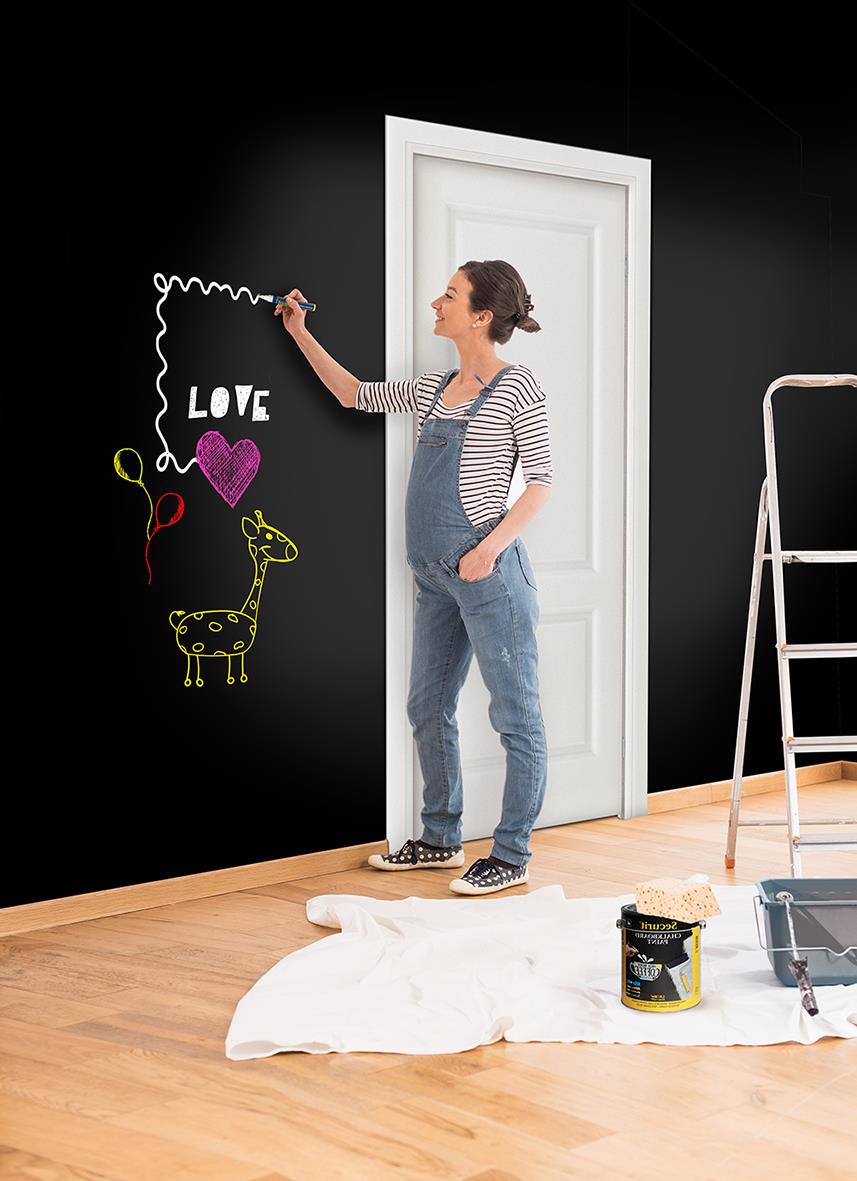 Kvinna målar med vit griffelfärg på en svart vägg, med en stege och målarutrustning i rummet