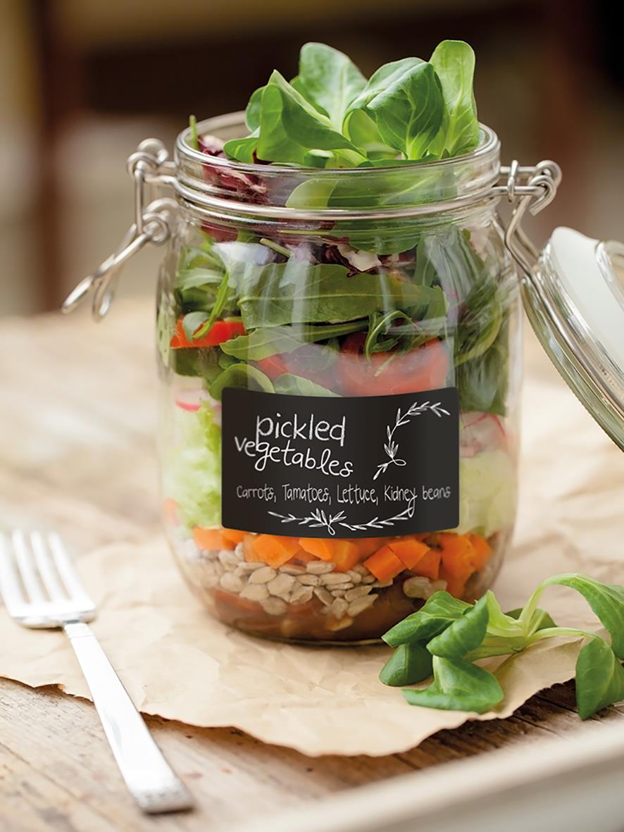 Glasburk med lager av färska grönsaker och en svart griffelklistermärke med text om picklade grönsaker
