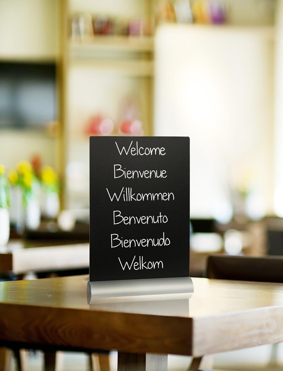 Svart griffeltavla med välkomstmeddelanden på flera språk, placerad på ett bord i ett rum