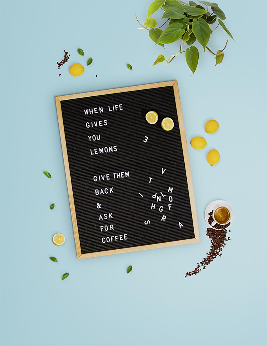 Svart bokstavstavla med citat, omringad av citroner, kaffe och gröna växter på ljusblå bakgrund