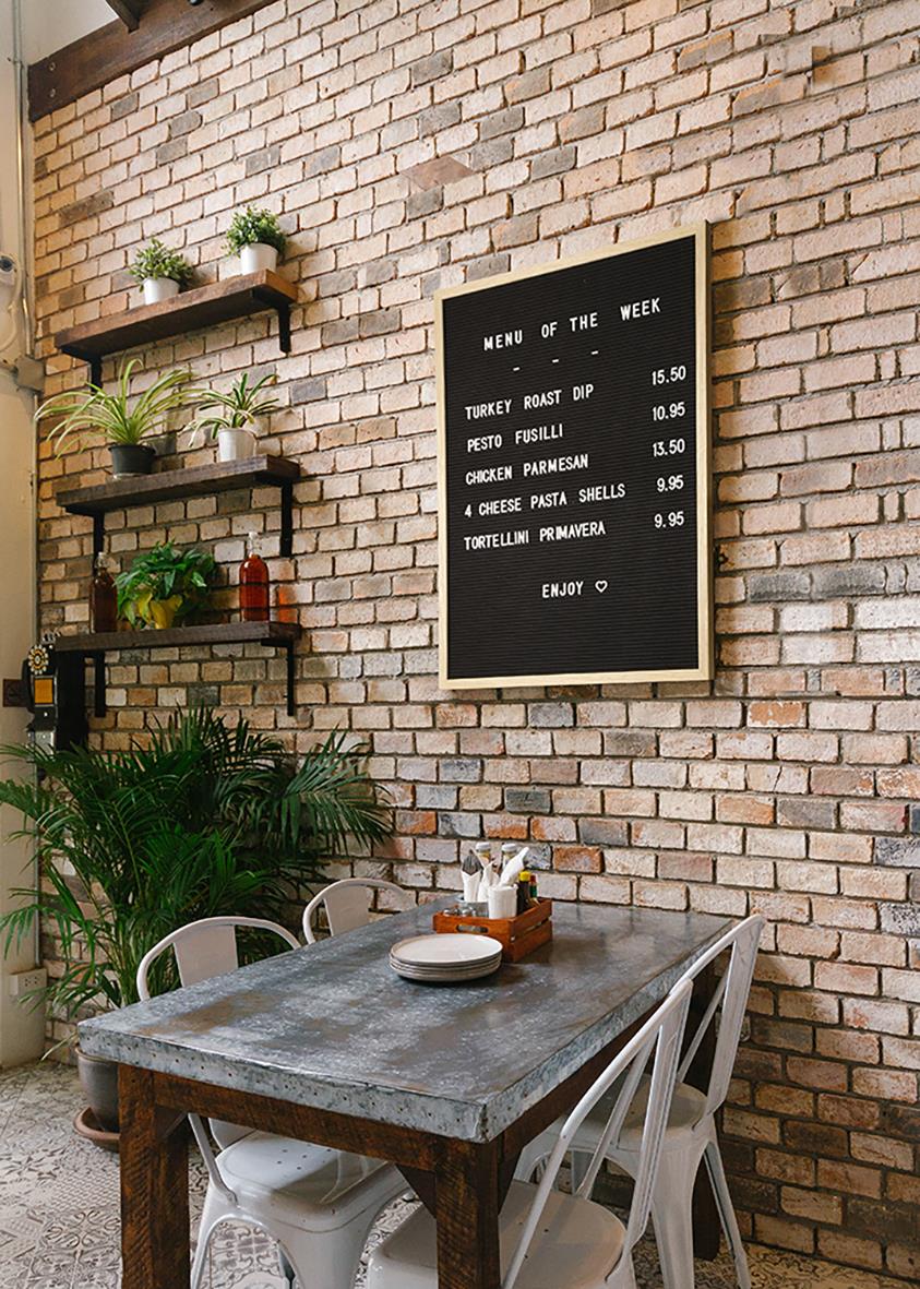 Inomhusbild av ett rustikt kök eller café med en brickbord, vita stolar, vägg med exponerad tegel, växt och en menybricka på väggen