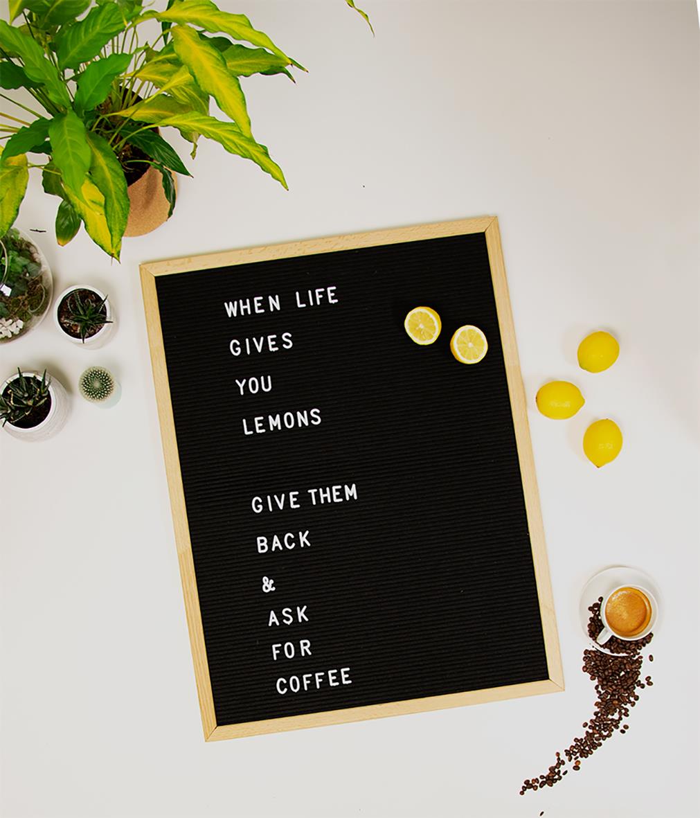 Svart bokstavstavla med citatet When life gives you lemons, give them back & ask for coffee, omgiven av växter och citroner