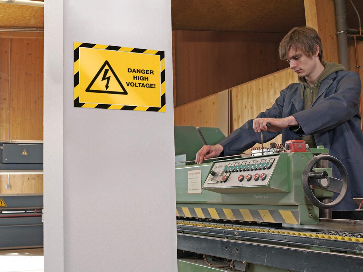 Person arbetar med maskin bredvid varningsskylt för hög spänning, gul och svart säkerhetsskylt