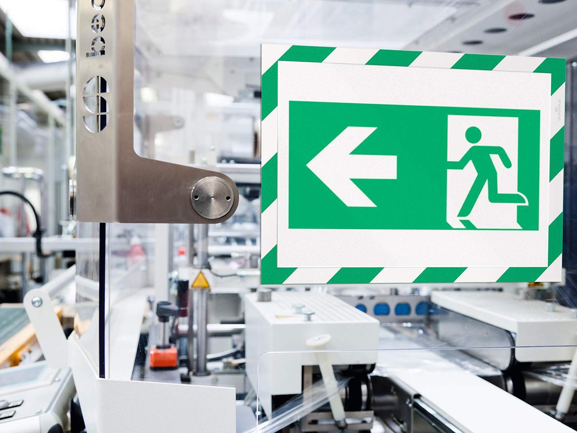 Grön/vit A4 säkerhetsskylt med pil och person som går ut genom en dörr i ett industriellt miljö