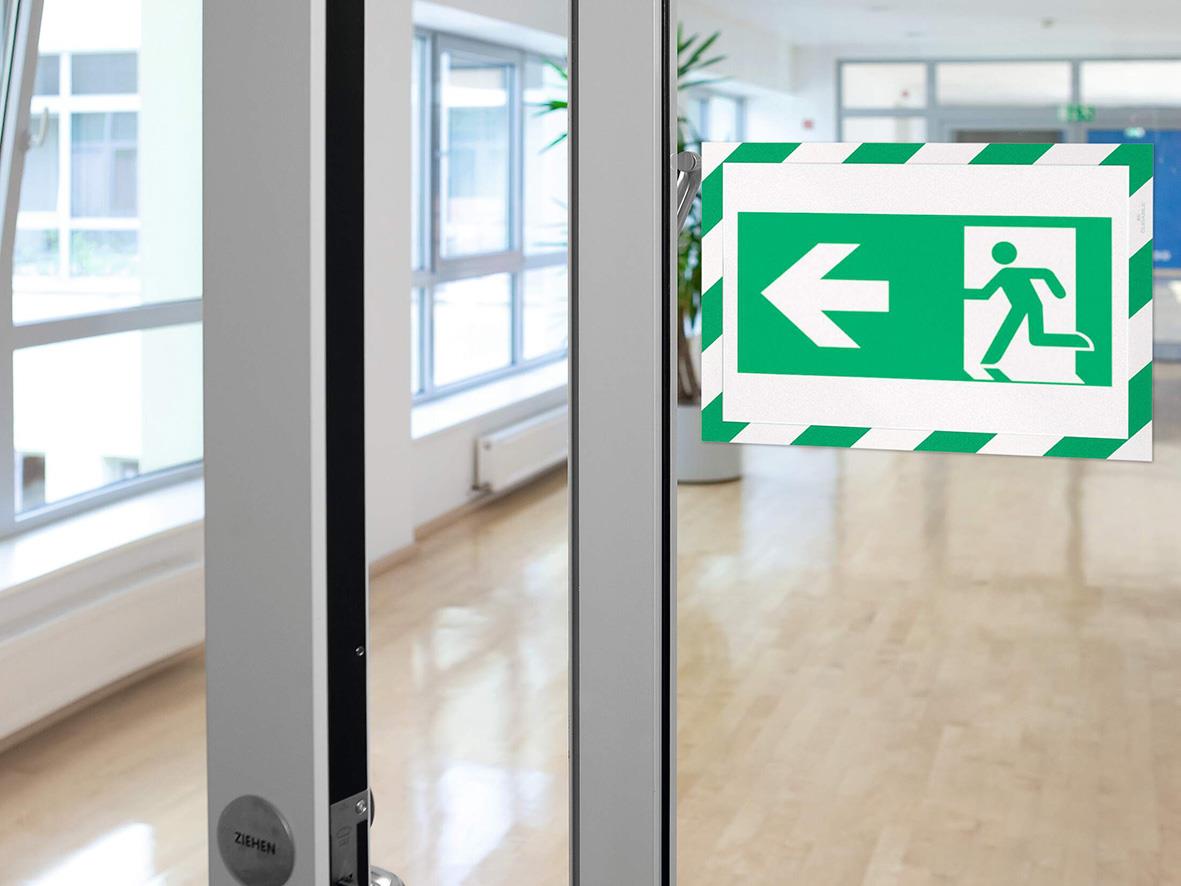 Grön/vit självhäftande säkerhetsskylt med pil och utrymningssymbol på glasdörr