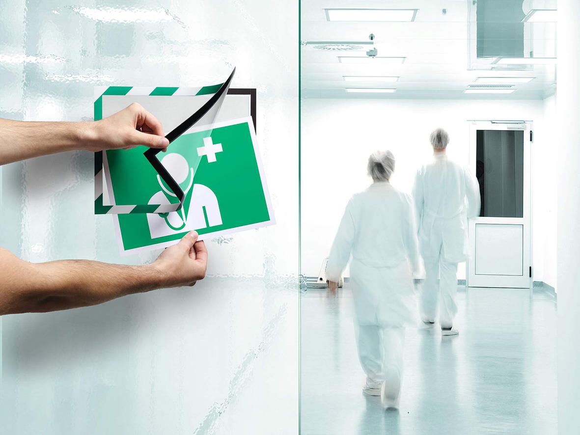 Person som sätter upp en självhäftande säkerhetsram i grönt och vitt på en vägg i ett sjukhus eller klinikmiljö
