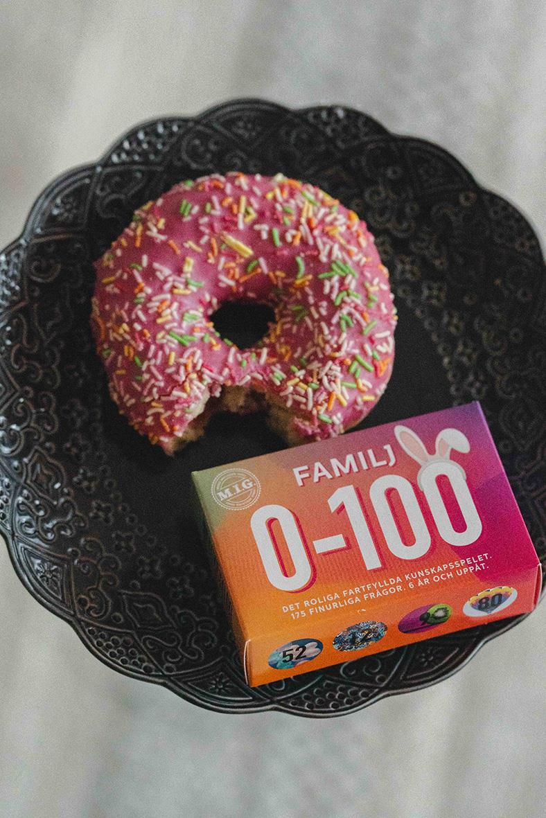 En rosa-glaserad donut med strössel på en svart tallrik bredvid ett spelpaket för familjequiz 0-100