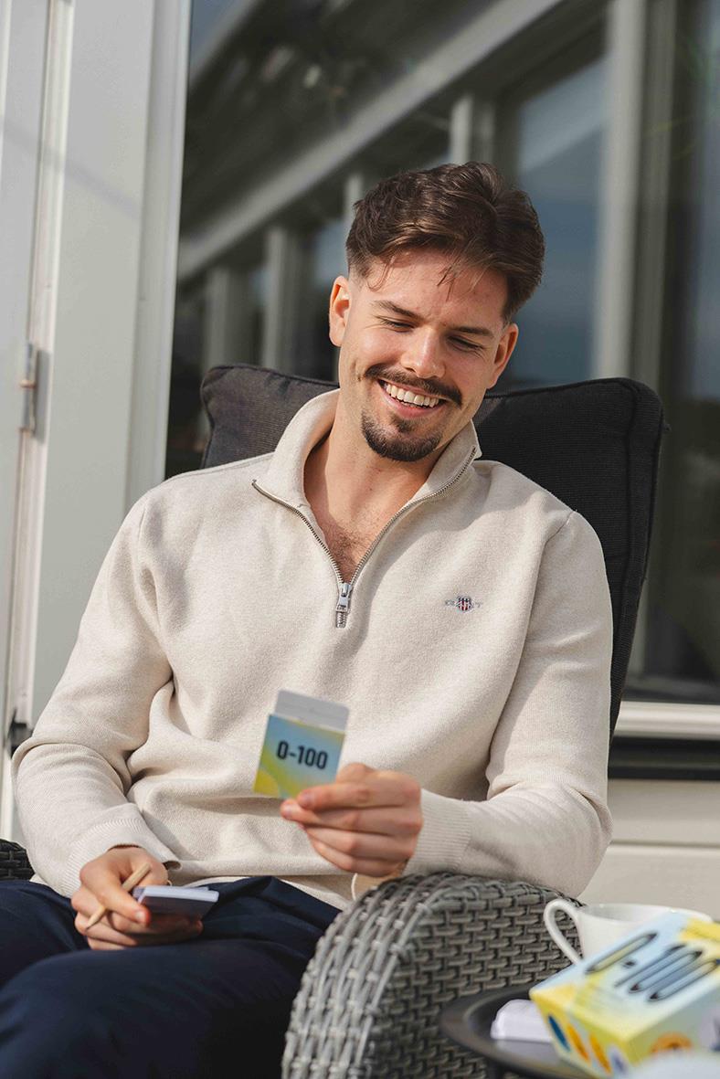 Ung man som spelar ett spel med kort, sitter avslappnad på en uteplats med glasväggar och koppar i bakgrunden