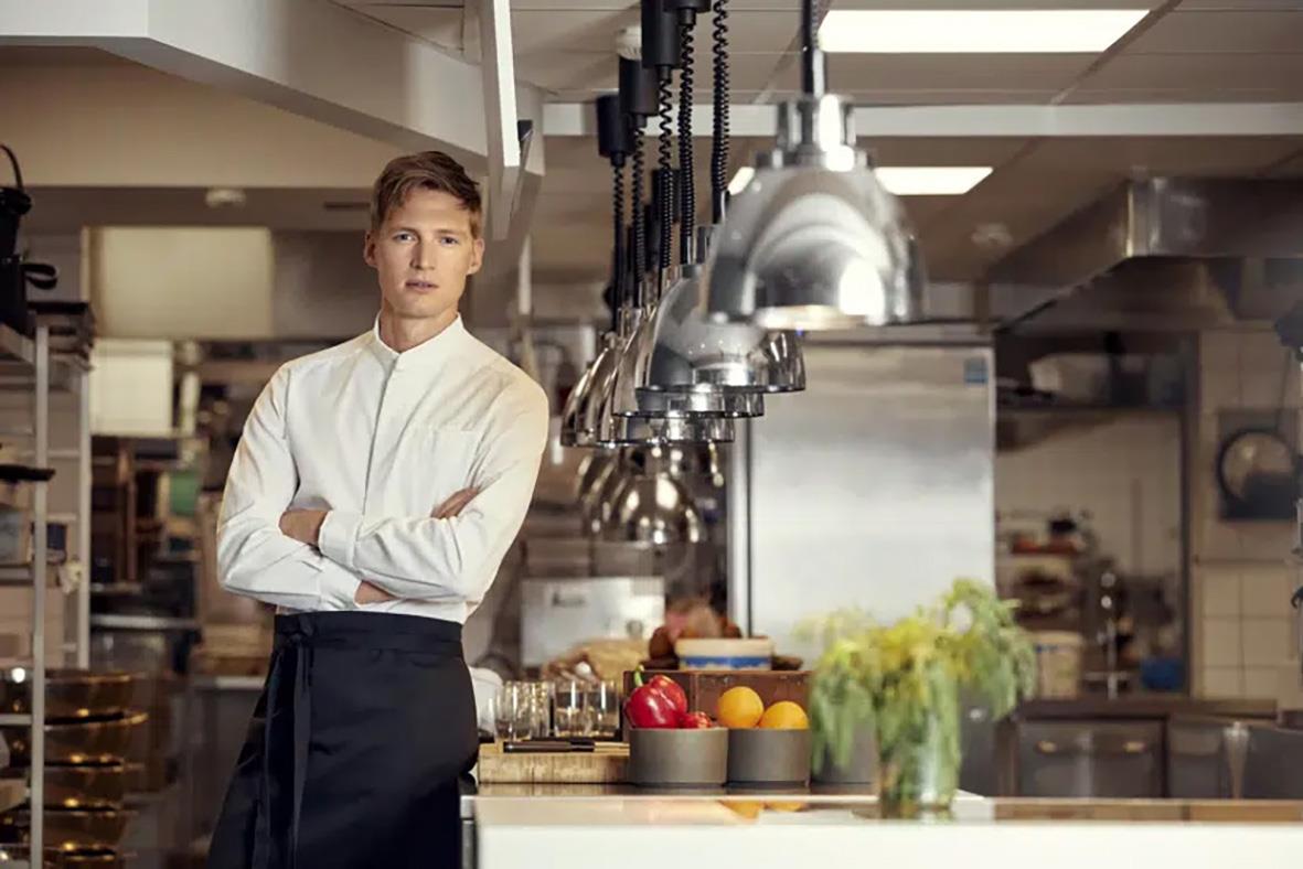 Kock i vit kockjacka står i ett professionellt kök med köksutrustning och ingredienser runt omkring