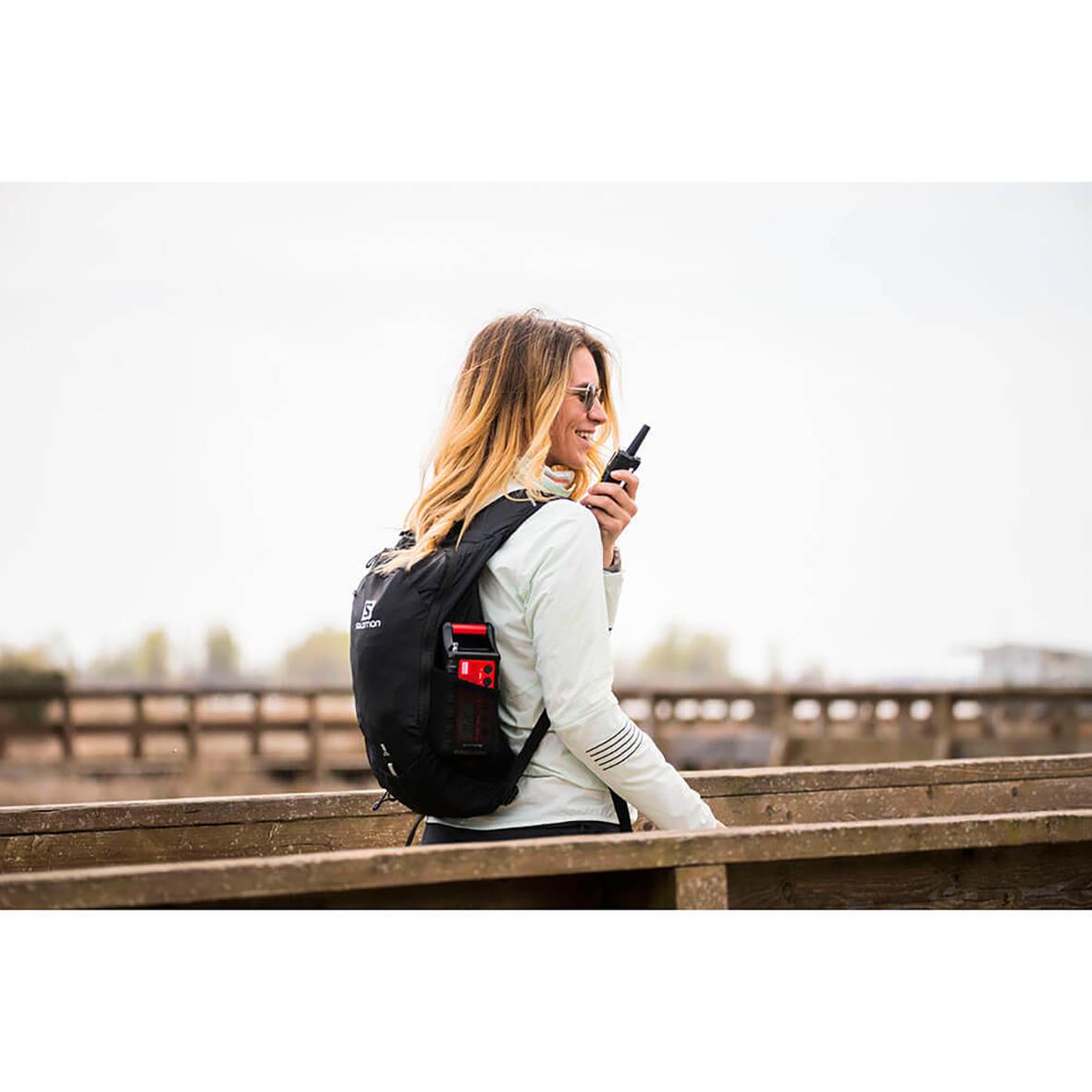 Kvinna med svart och röd walkie talkie Midland XT10 Pro, utomhus på bro, bär ryggsäck och pratar i radion