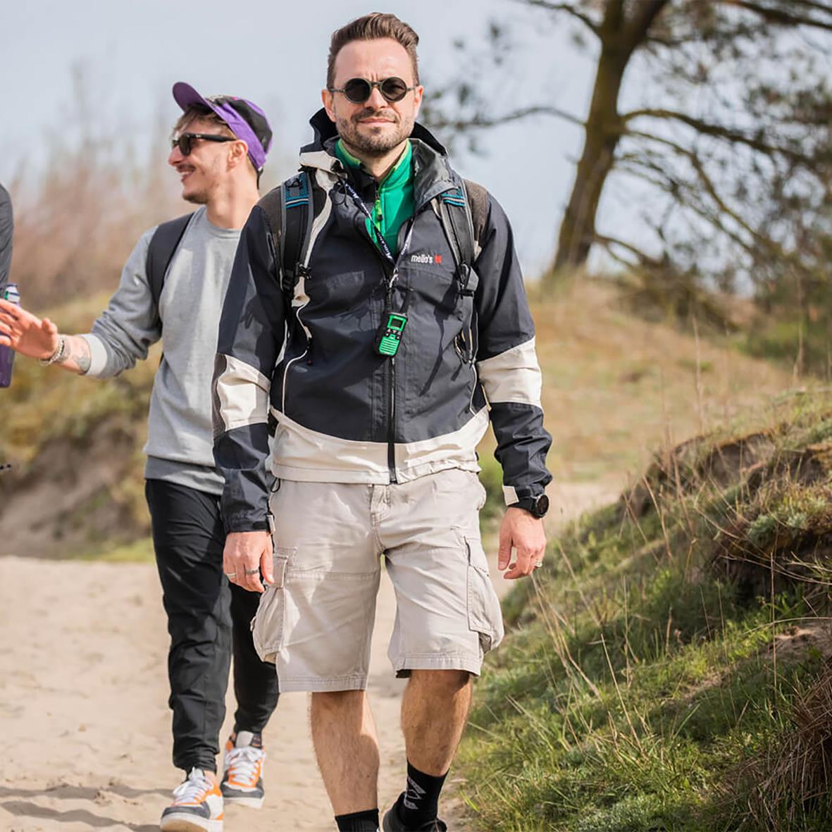 " personer vandrar utomhus, en med ryggsäck och walkie talkie, i en naturmiljö med träd och gräs"