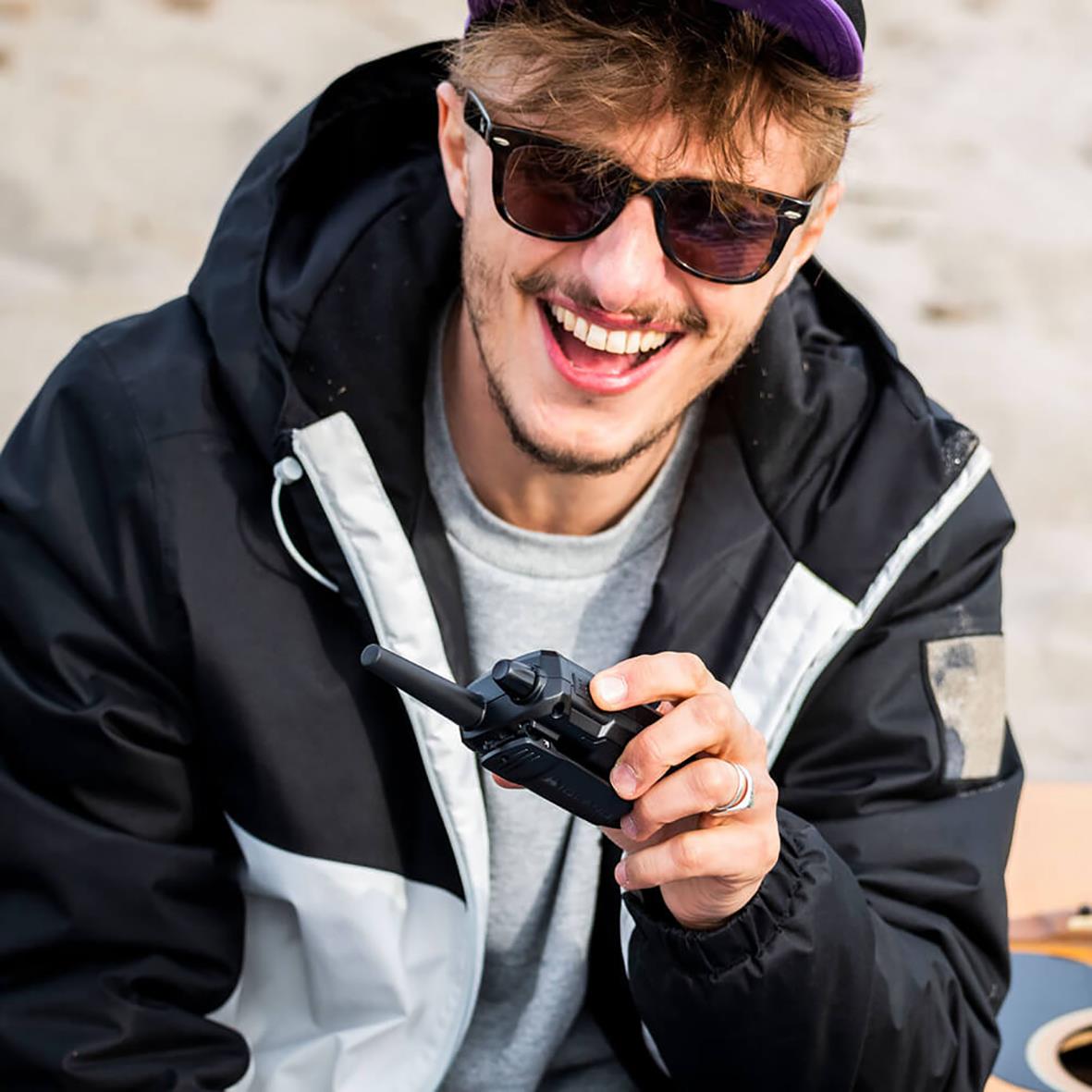 Ung man med solglasögon och svart/mörkgrön jacka håller en walkie talkie och ler