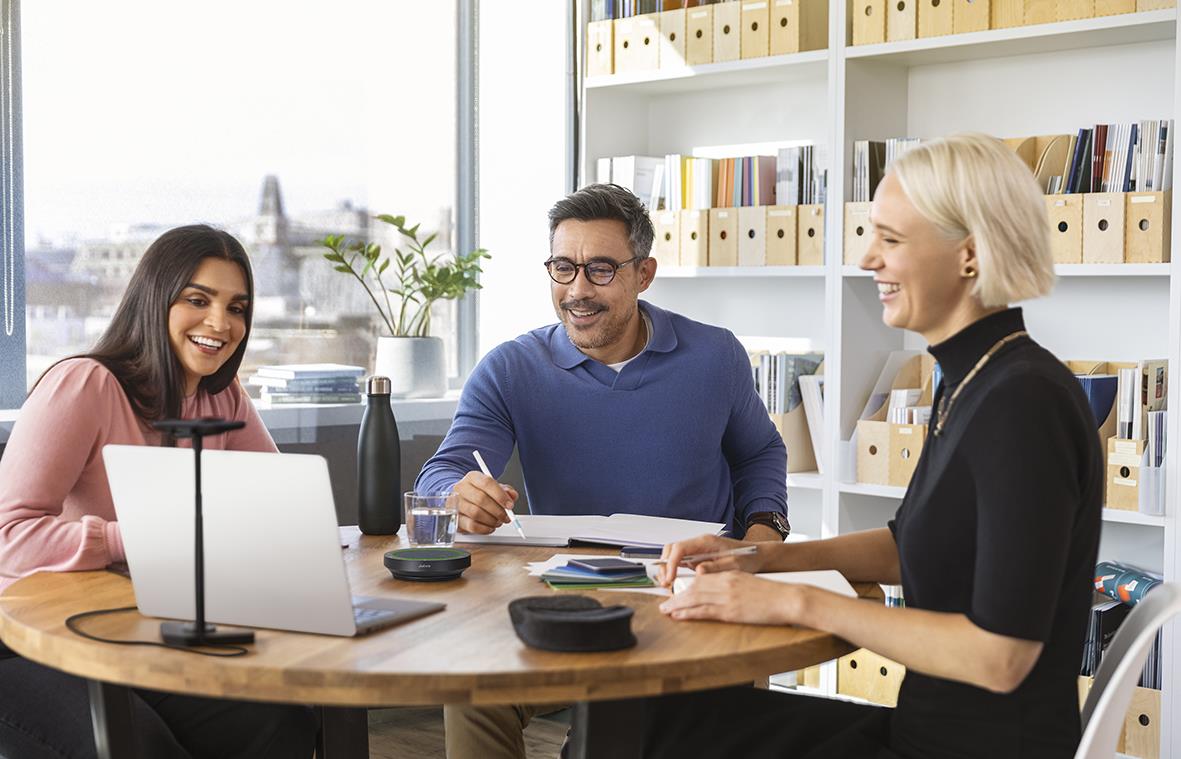 Tre personer i ett mötesrum med laptop och böcker, diskuterar och skrattar