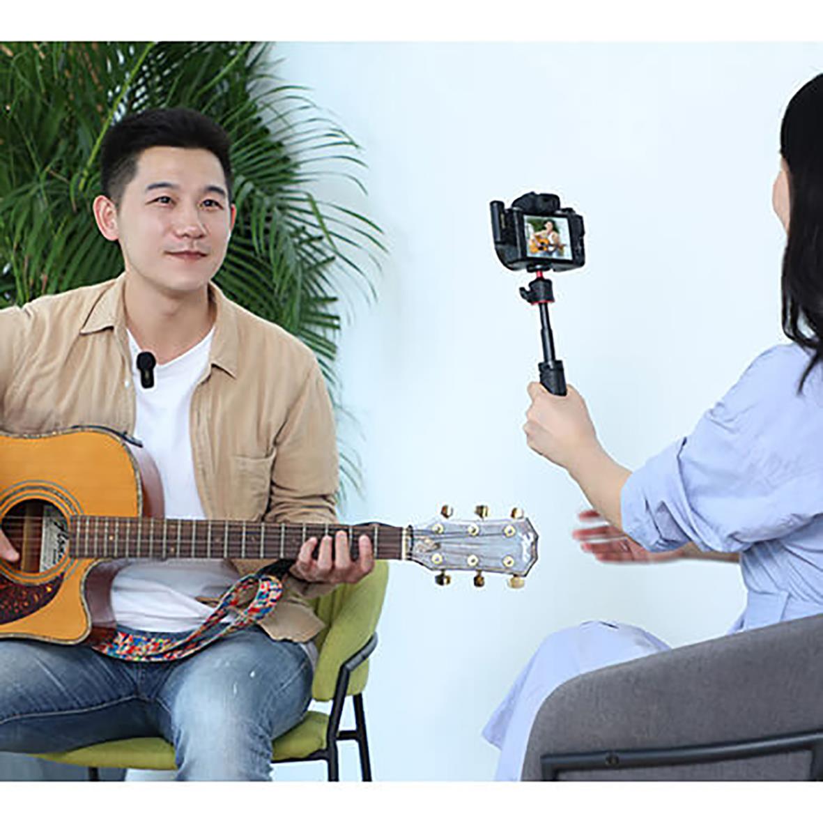 Man spelar gitarr medan en annan person filmar med en videokamera, i en inomhusmiljö med gröna växter i bakgrunden