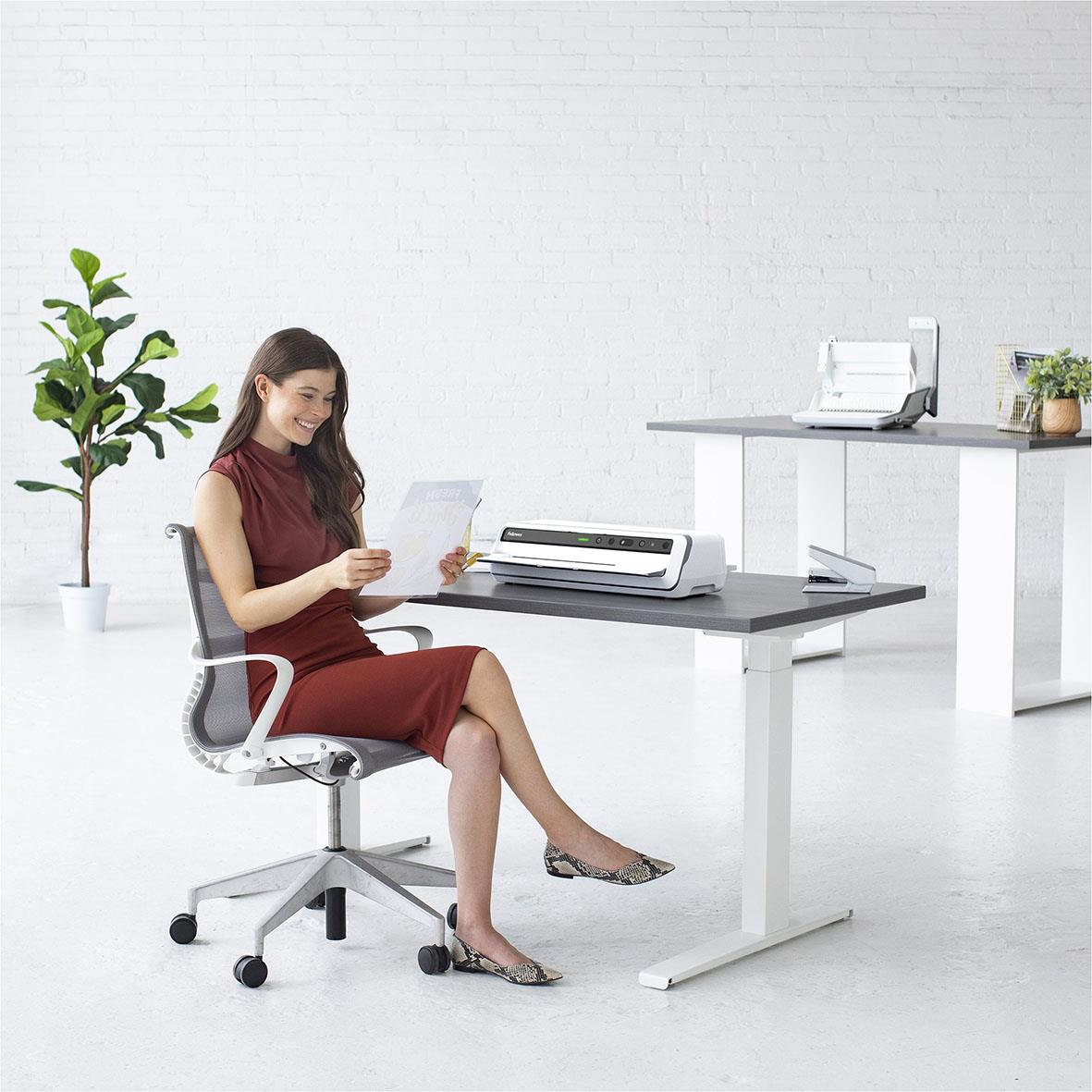 Kvinna sitter vid skrivbord med lamineringsmaskin Fellowes Venus A3, vitt rum, grönt växt i bakgrunden