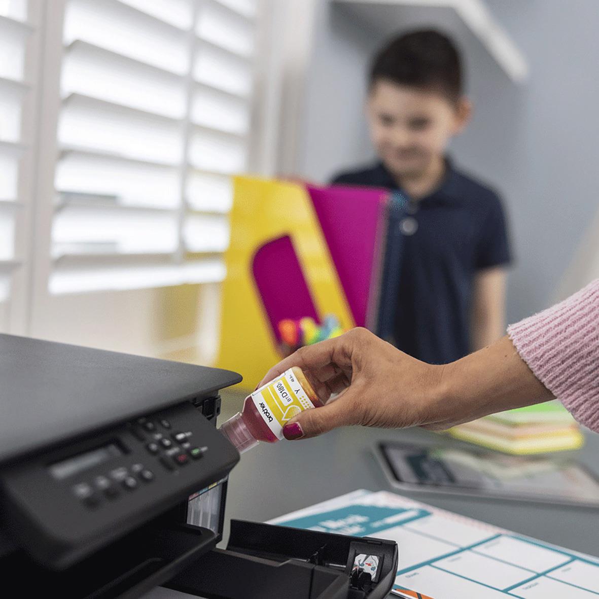 Person fyller på bläck i en multifunktionsskrivare Brother DCP-T580DW, med barn i bakgrunden