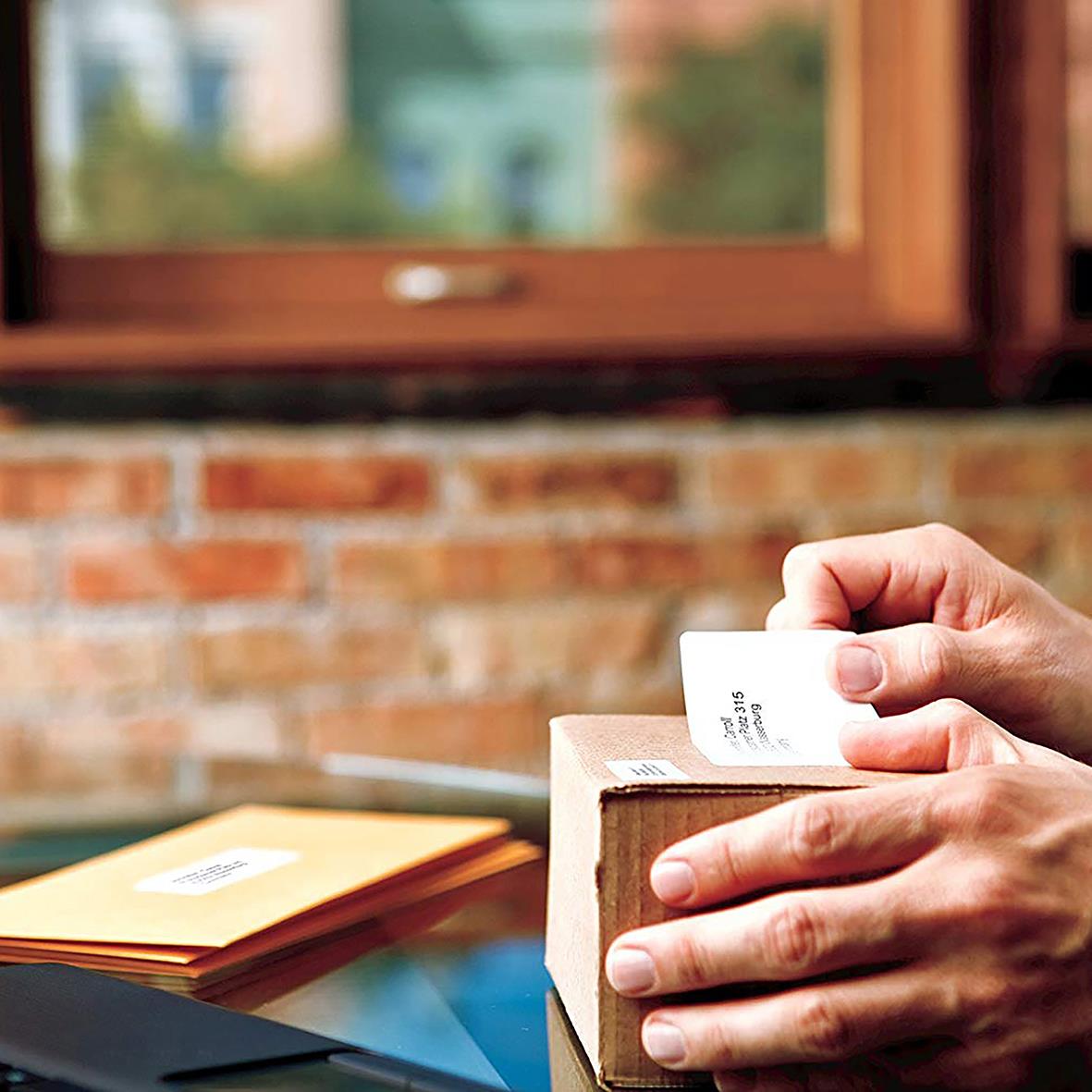 Person skriver ut etiketter med Dymo Labelwriter på ett träbord, bakgrund av fönster och tegelvägg