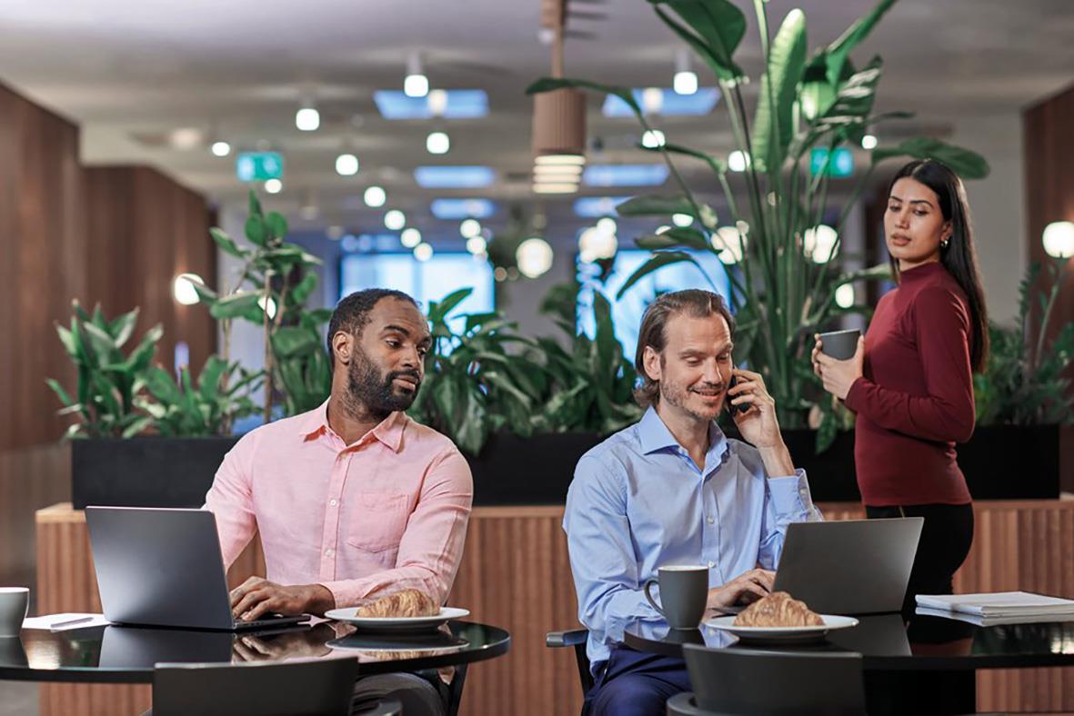 Två män vid ett bord med laptops och en kvinna som står med en kopp i en modern kontorsmiljö med gröna växter
