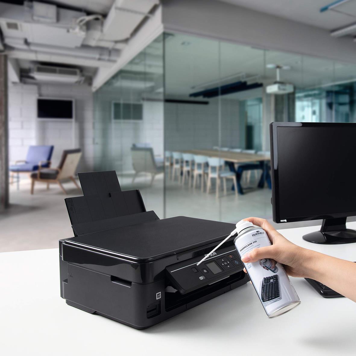 Person använder tryckluftsspray för att rengöra en skrivare i ett modernt kontorsutrymme