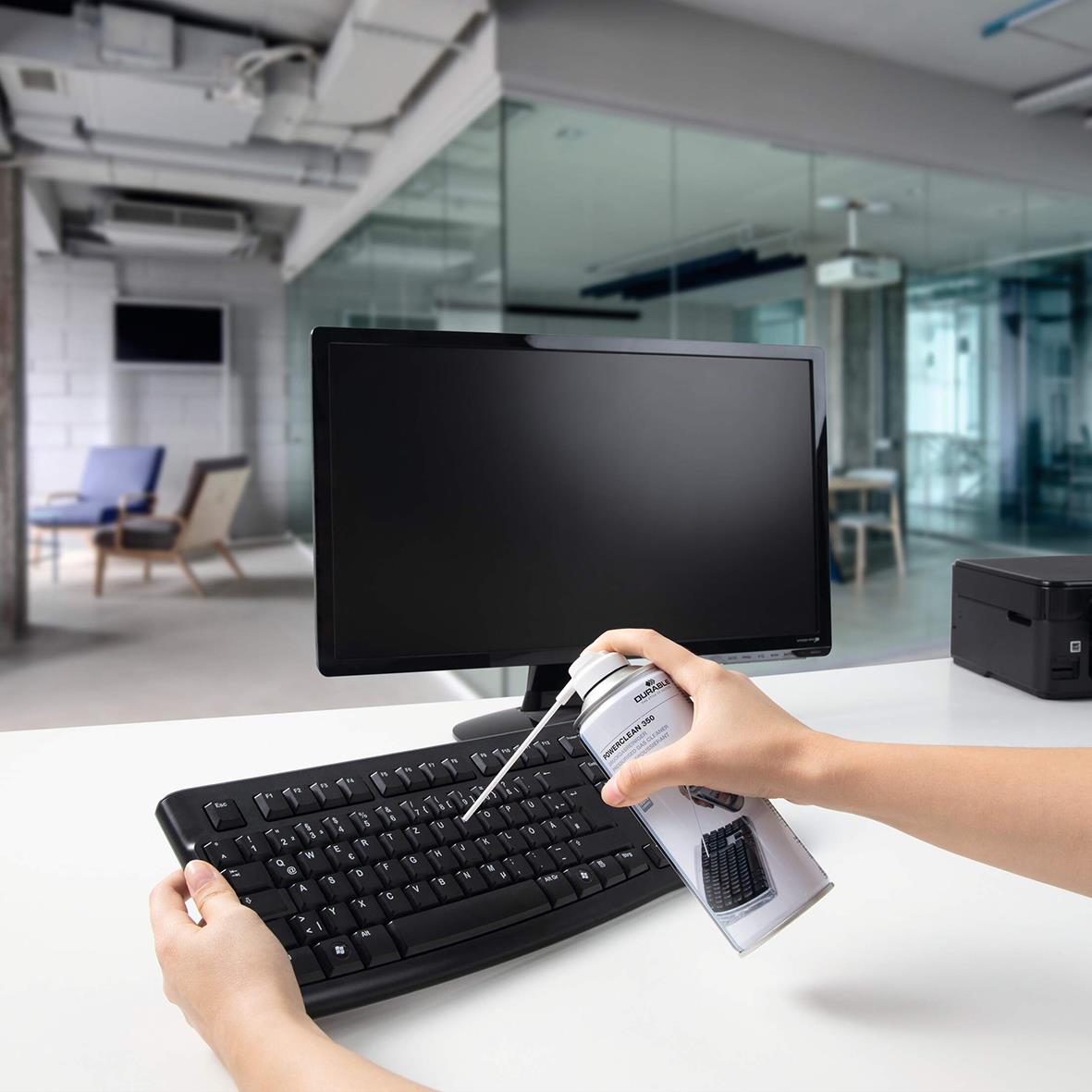Person använder tryckluftsspray för att rengöra en datorskärm och tangentbord i ett modernt kontorsutrymme