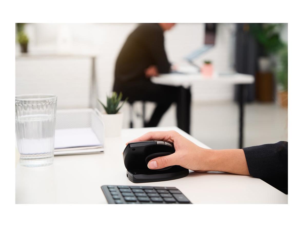 Hand som använder ergonomisk mus på skrivbord med dator och växt i bakgrunden