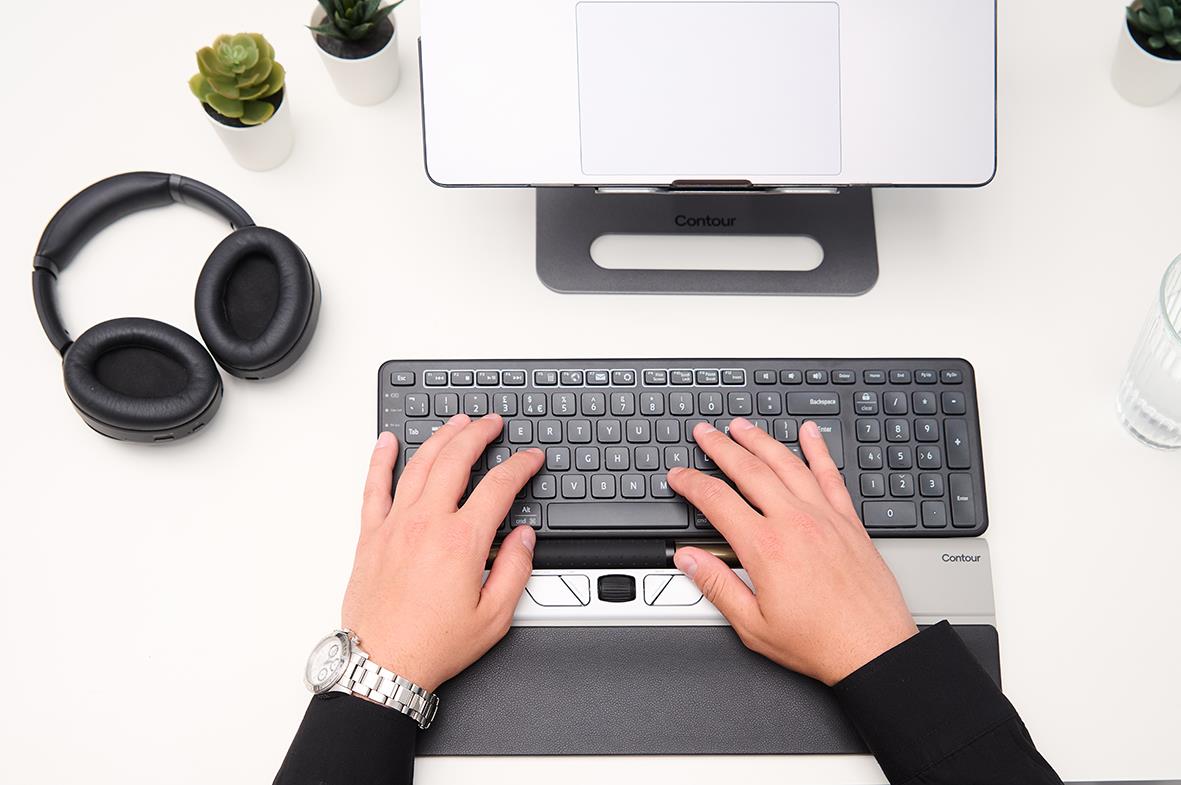 Person som använder en trådlös ergonomisk mus och tangentbord på skrivbord med laptop och växter