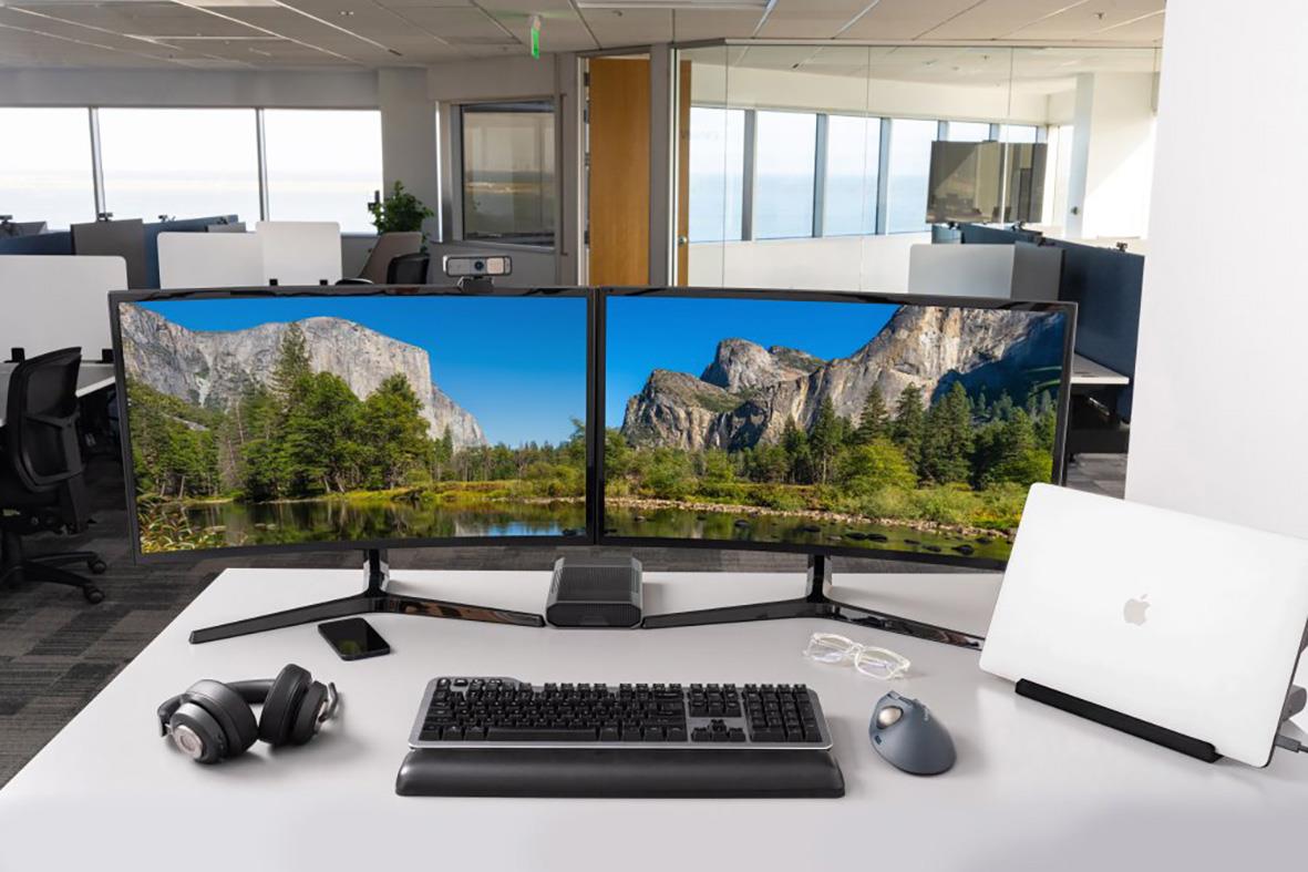 Två datorskärmar med landskapsbild, tangentbord, mus, hörlurar och laptop på skrivbord i kontorsmiljö