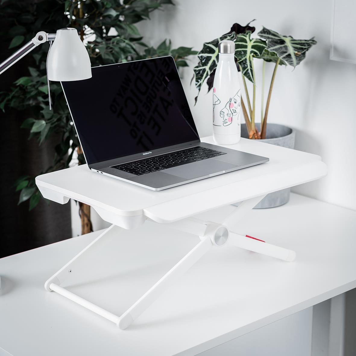 Portabelt höj- och sänkbart ståbord med laptop, lampa och vattenflaska på ett vitt skrivbord