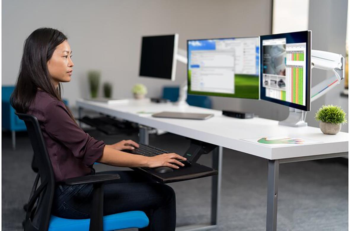 Kvinna vid skrivbord med dubbla monitorarmar och två datorskärmar i ett kontorsmiljö