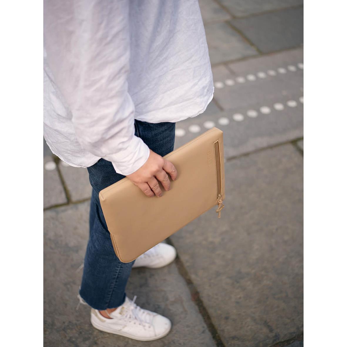 Person håller ett beige neopren datorfodral för 13 tum med gulddragkedja på en stadsgata