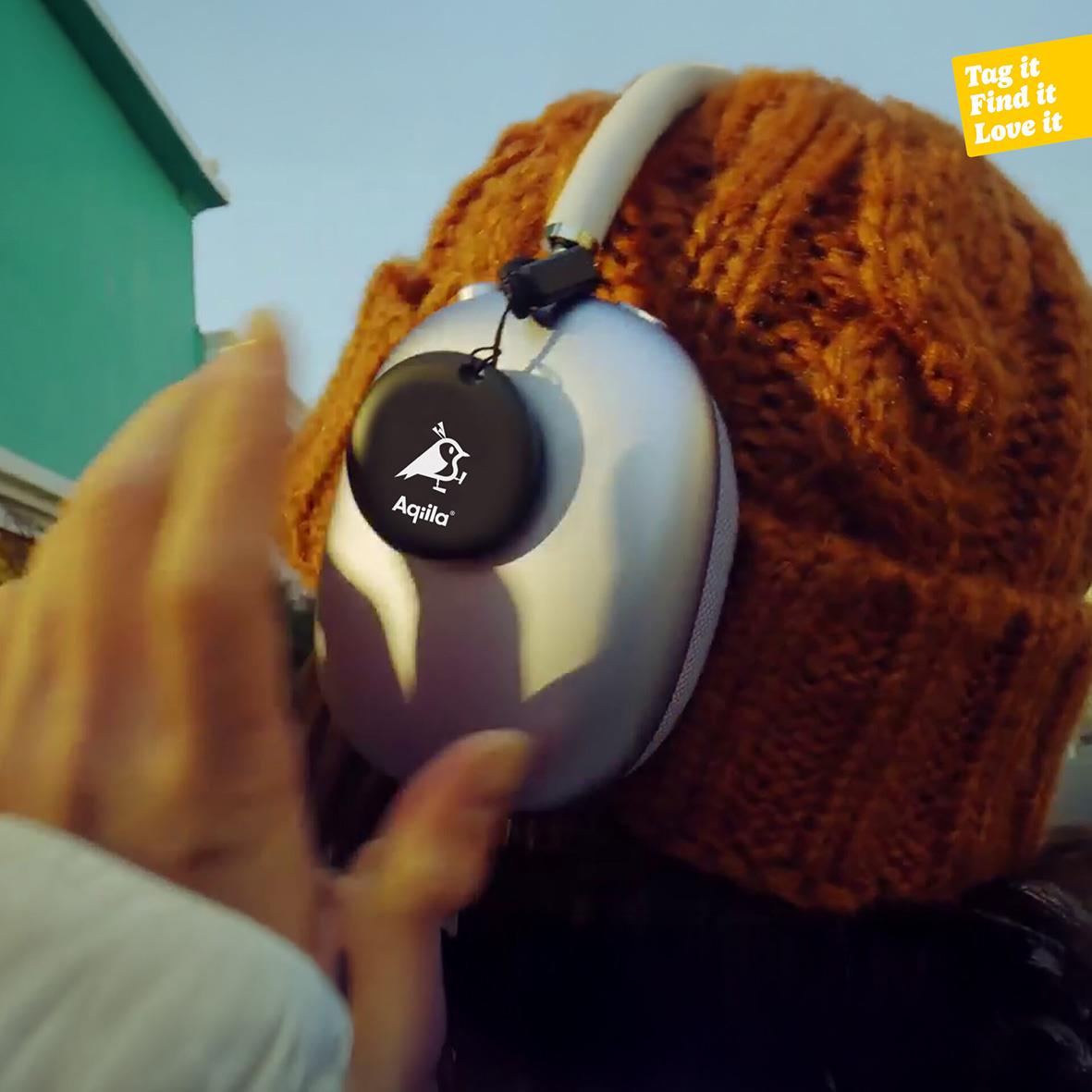 Person bär hörlurar med Aqiila Tracker Tagbird på huvudet, orange mössa och grön byggnad i bakgrunden