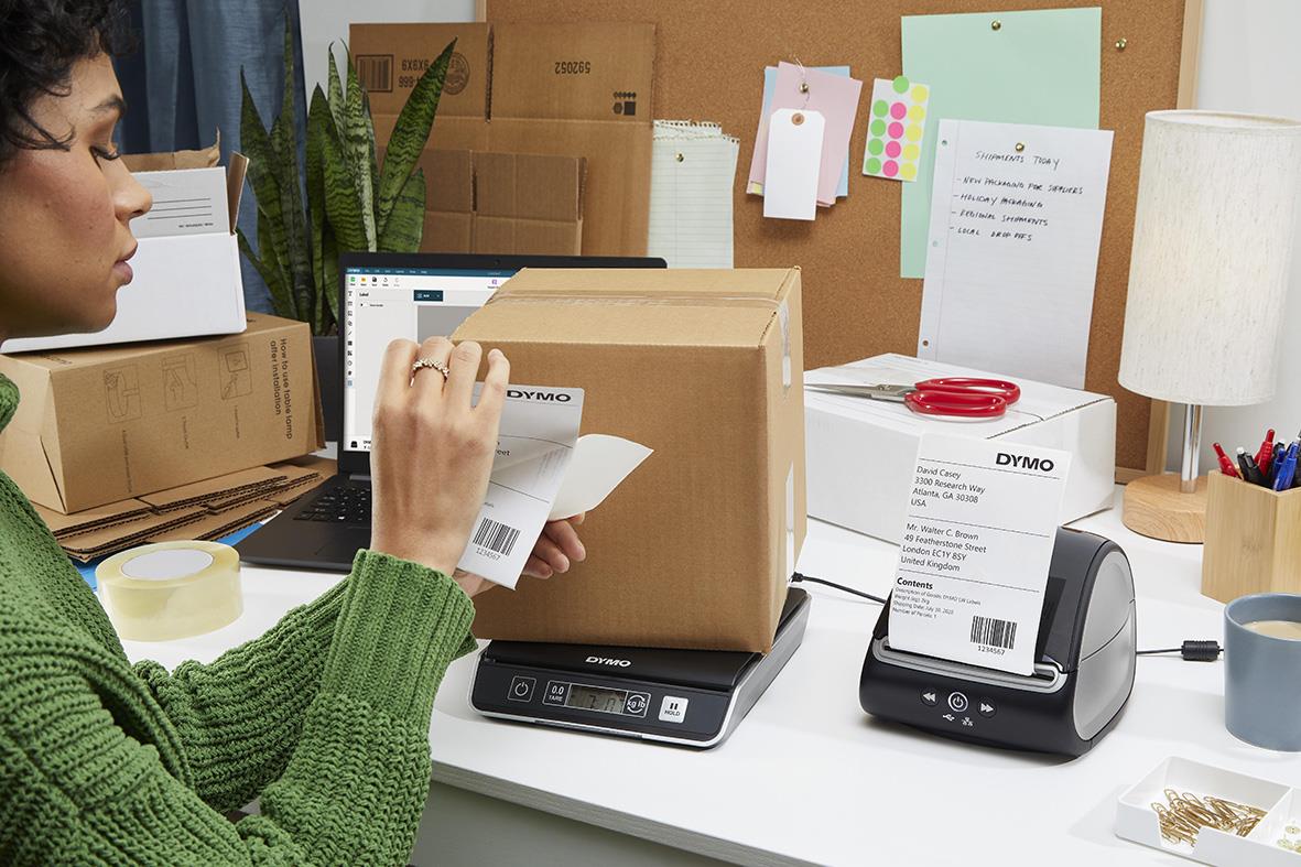 Person väger ett paket på en Dymo M2 2kg våg i ett kontorsmiljö