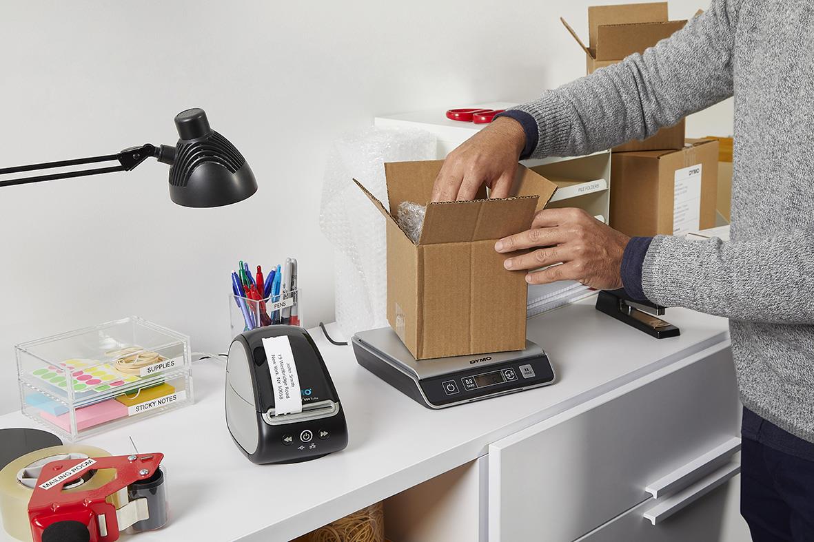 Person väger ett paket på en digital våg med USB-anslutning och 10 kg kapacitet i ett kontors- eller lagerutrymme