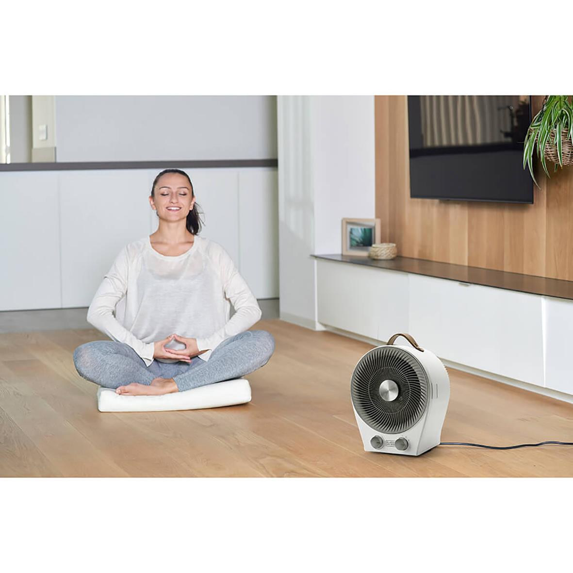 Kvinna i meditationställning framför en svart och vit värmefläkt Black+Decker 2IN1 i ett modernt vardagsrum