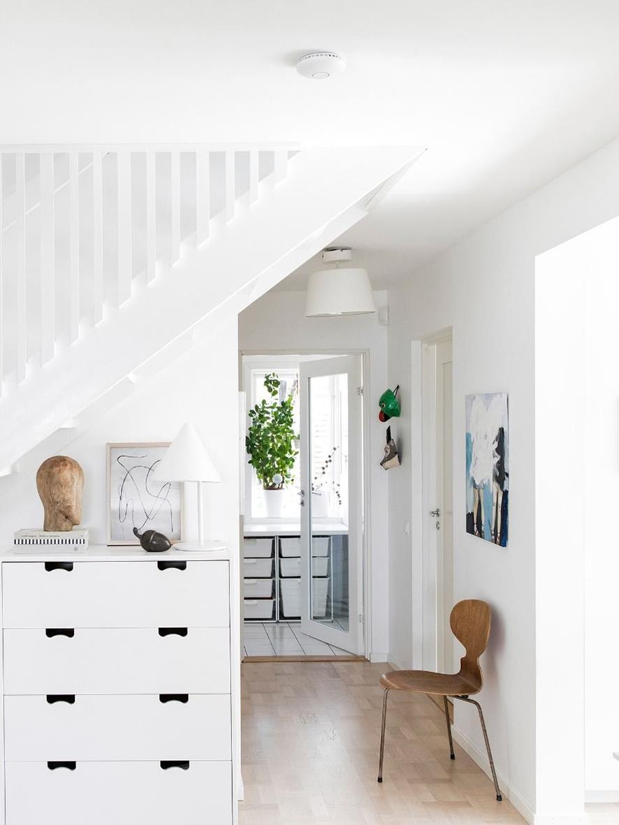 Vitt rum med trapp, vit byrå, konstverk, stol och dörr med glas, växt och väggdekoration, ljus och luftigt.