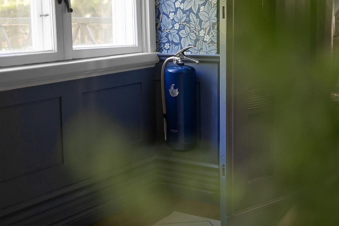 Blå och silver brandsläckare Housegard Pulver 6kg placerad i ett hörn inomhus