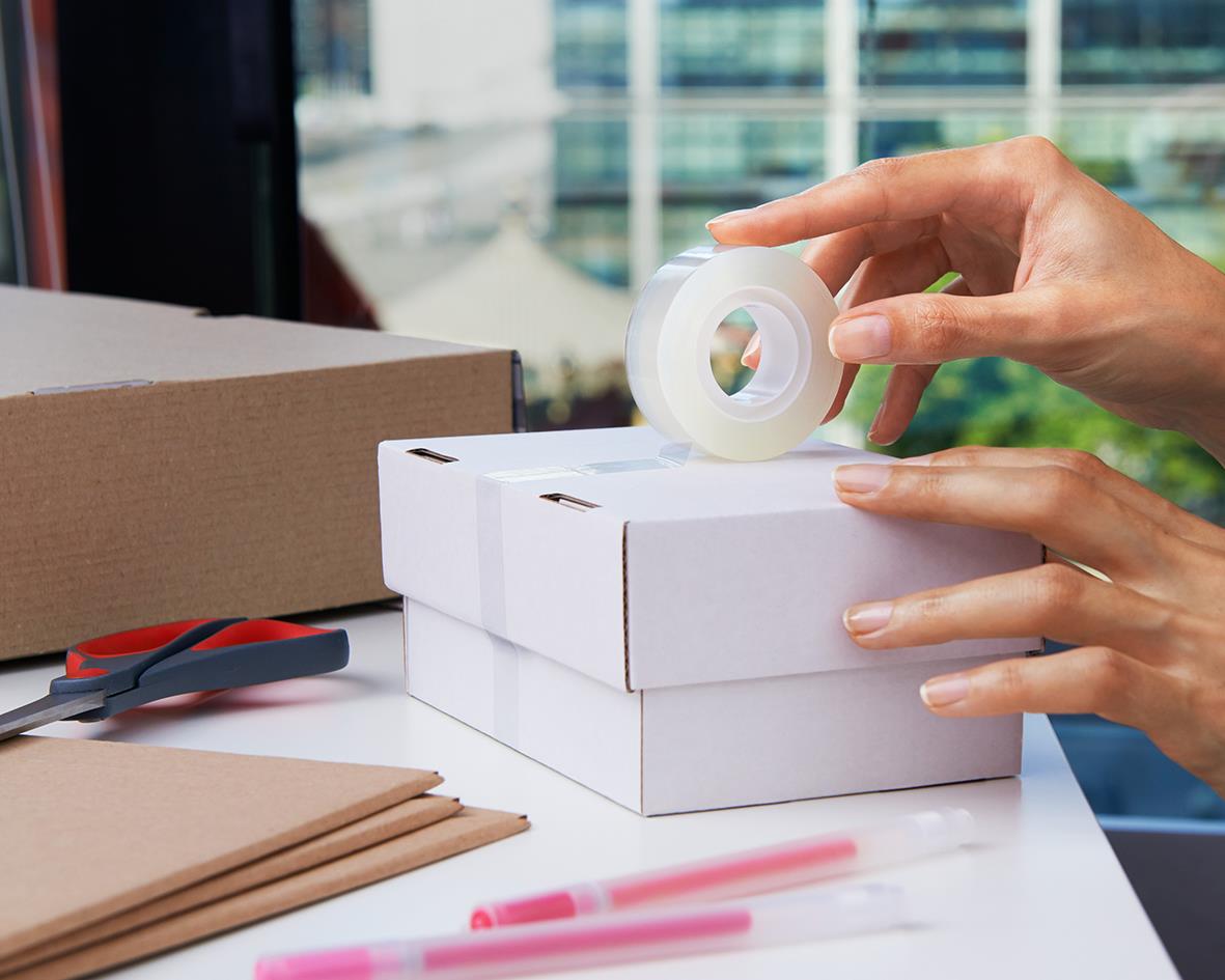 Person som använder Scotch 600 Crystal kontorstejp för att täcka en kartong på ett skrivbord med kontorsmaterial