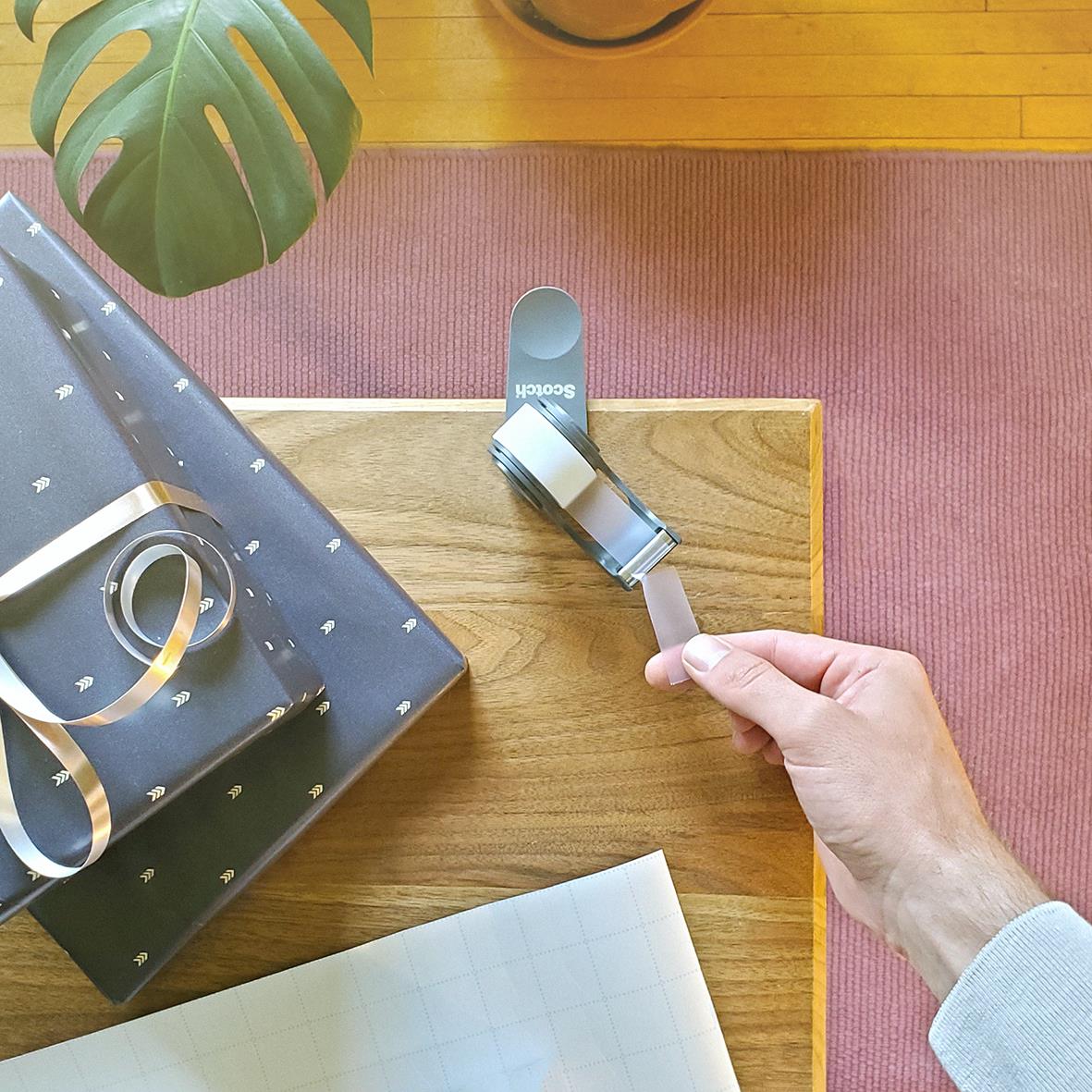 Tejhållare Scotch C19 Clip & Twist Grå 33m hålls i hand över bord med presentförpackning och blad i bakgrunden