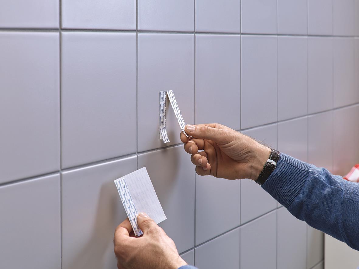 Person använder självhäftande Tesa Powerstrips för att fästa något på kakelvägg