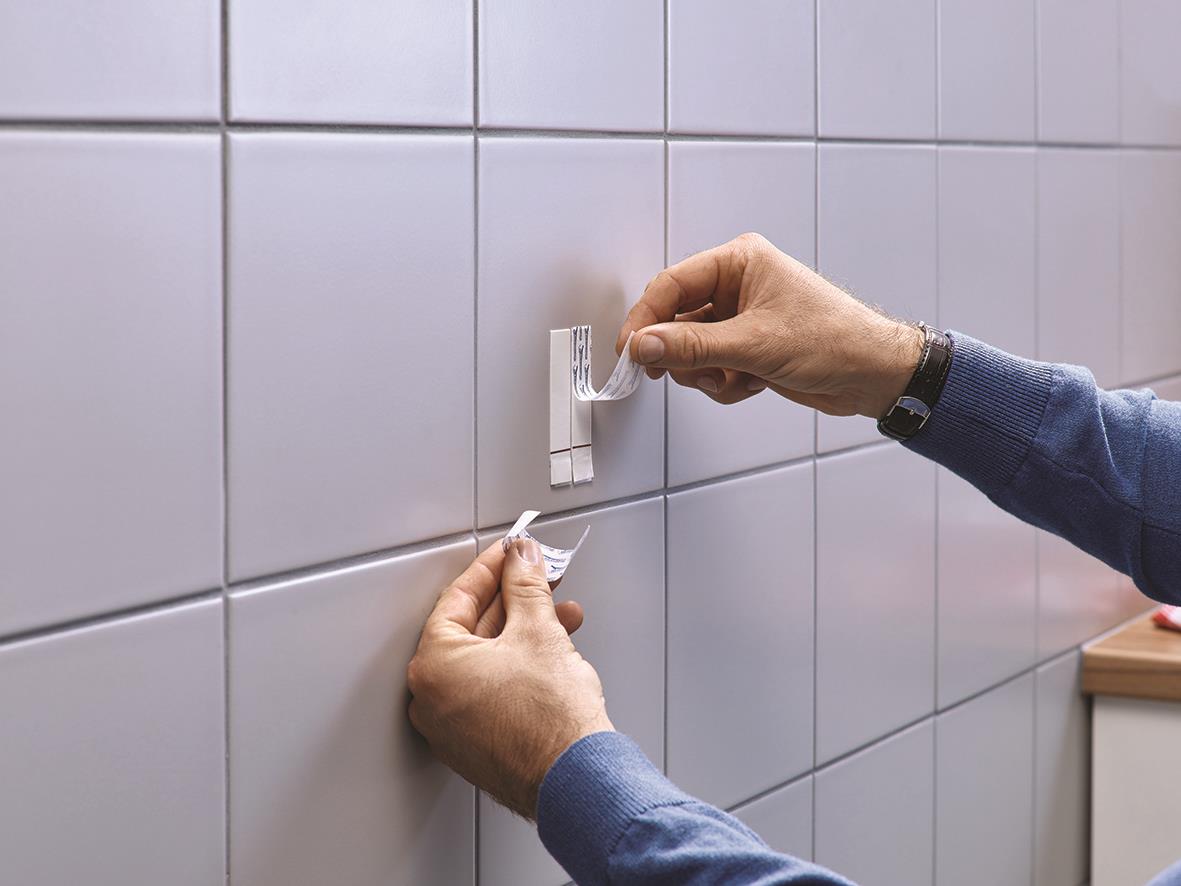 Person applicerar Tesa Powerstrips på kakelvägg för att fästa något, justerbar och klar för belastning upp till 4kg