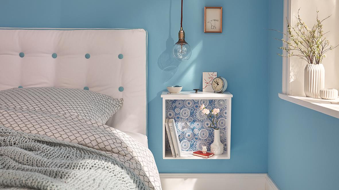 Sovrum med blå vägg, vit sänggavel, vägglampa och växt i fönster, samt en vit vägghylla med böcker och dekorationer